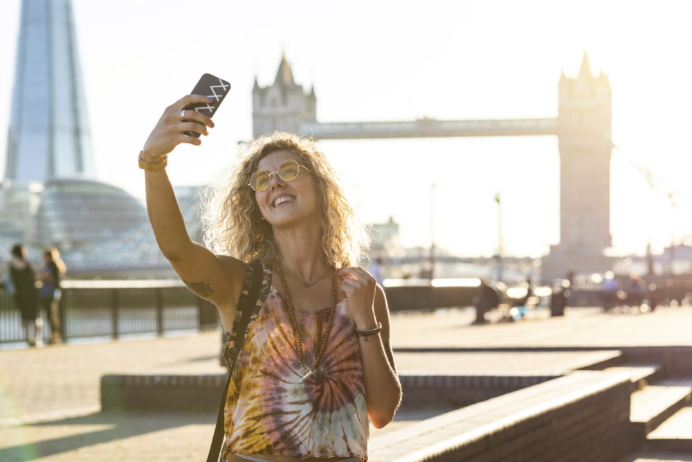 Kvinne på ferie tek ein selfie framfor Tower Bridge i London, smilande i solskinet med solbriller og fargerik topp.