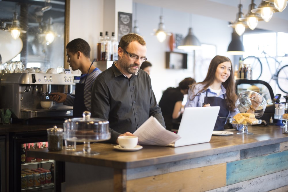 Mann les avisa medan han nyt ein kopp kaffi på ein kafé og har laptopen open.