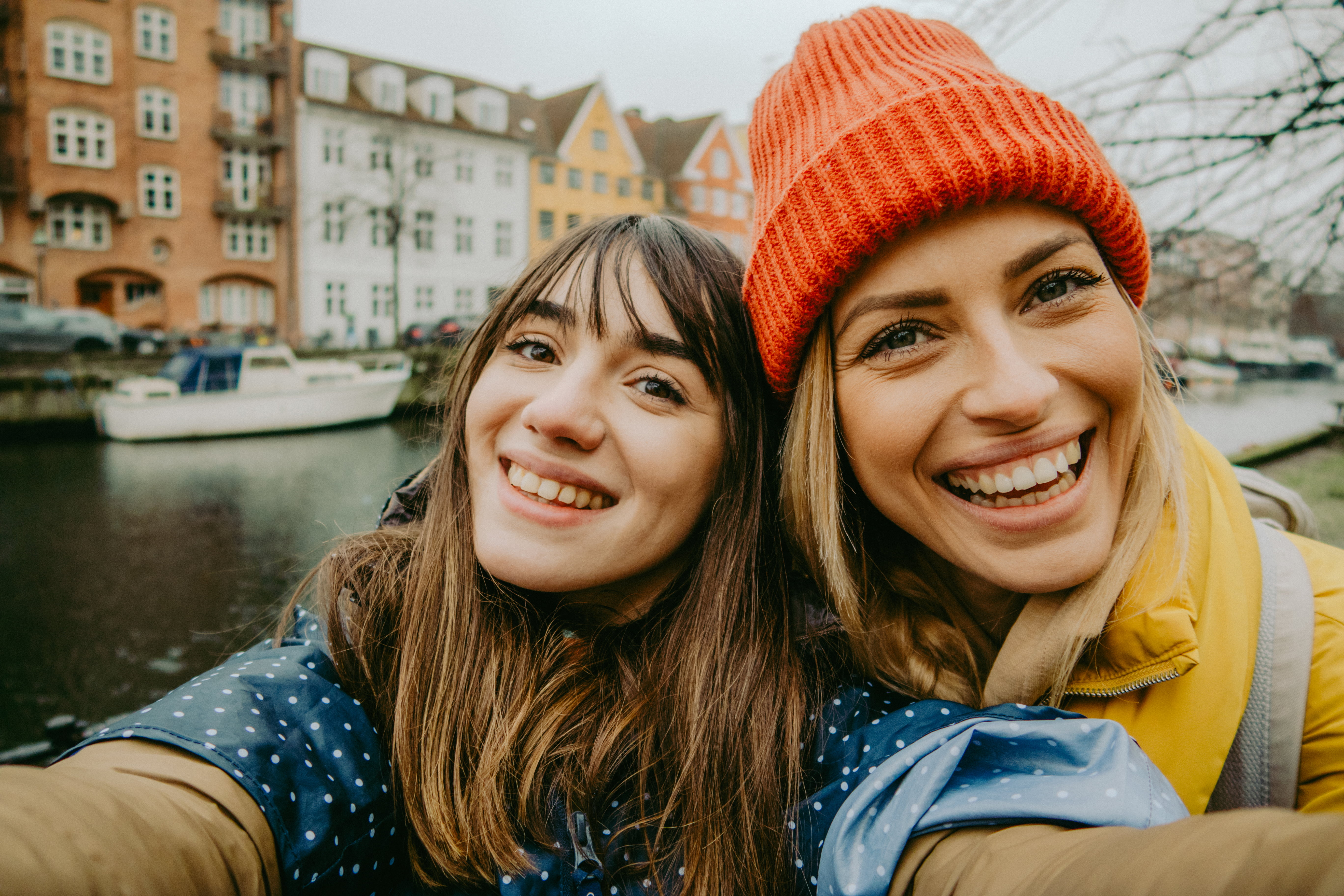 To smilande veninner tek ein selfie med ein pittoresk kanal og fargerike bygningar i bakgrunnen, som representerer glede og fellesskap i kvardagsaugneblikka.