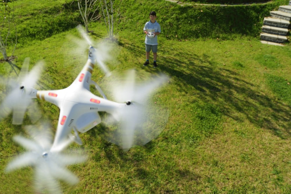 Ein ung gut styrer ein flygande kvit drone på ein solrik dag i ein frodig hage. Drona er i fokus med bevegelsesuskarpheit på propellane, og viser teknologien i bruk i eit naturleg og morosamt miljø.