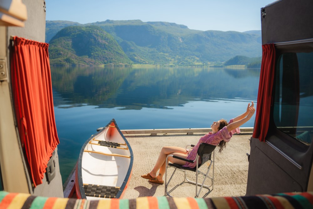Ei kvinne slappar av i ein samanleggbar stol ved sida av ei campingvogn, strekkjer armane lukkeleg mot sola. Framfor ho er ein kano plassert ved breidda av ein roleg fjord omgjeven av frodige fjell.