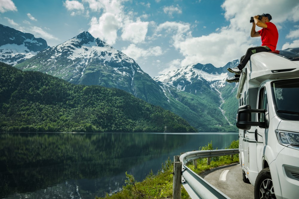 Ein person i raud t-skjorte står på taket av ein kvit bobil og bruker kikkert for å sjå på landskapet. Bakgrunnen viser ein spegelblank fjord omkransa av frodige grøne skogar og snødekte fjelltoppar.
