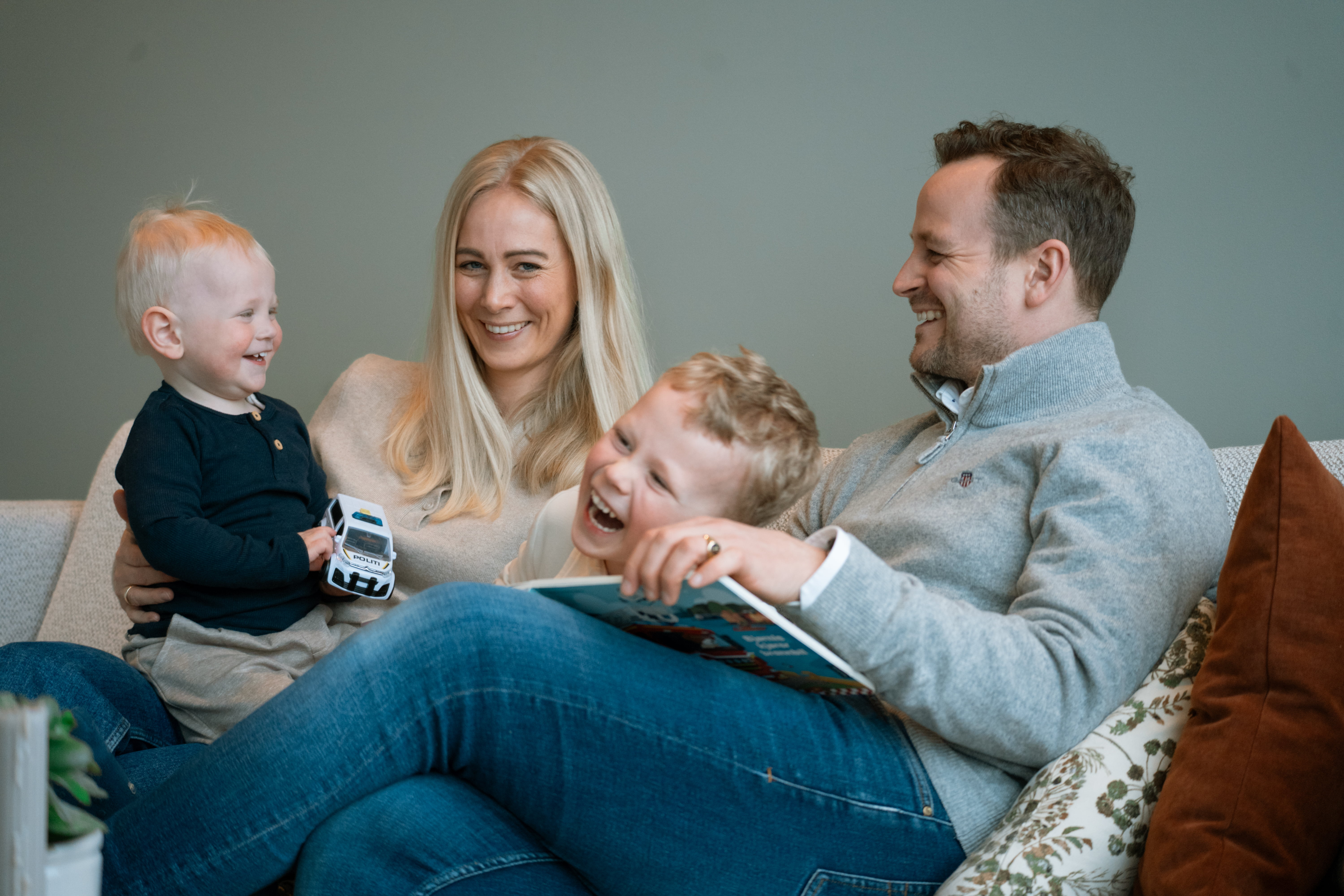 Familie sit i sofaen og les ei bok saman. Eitt barn held ein leikebil, og det er puter og grøne plantar i rommet.