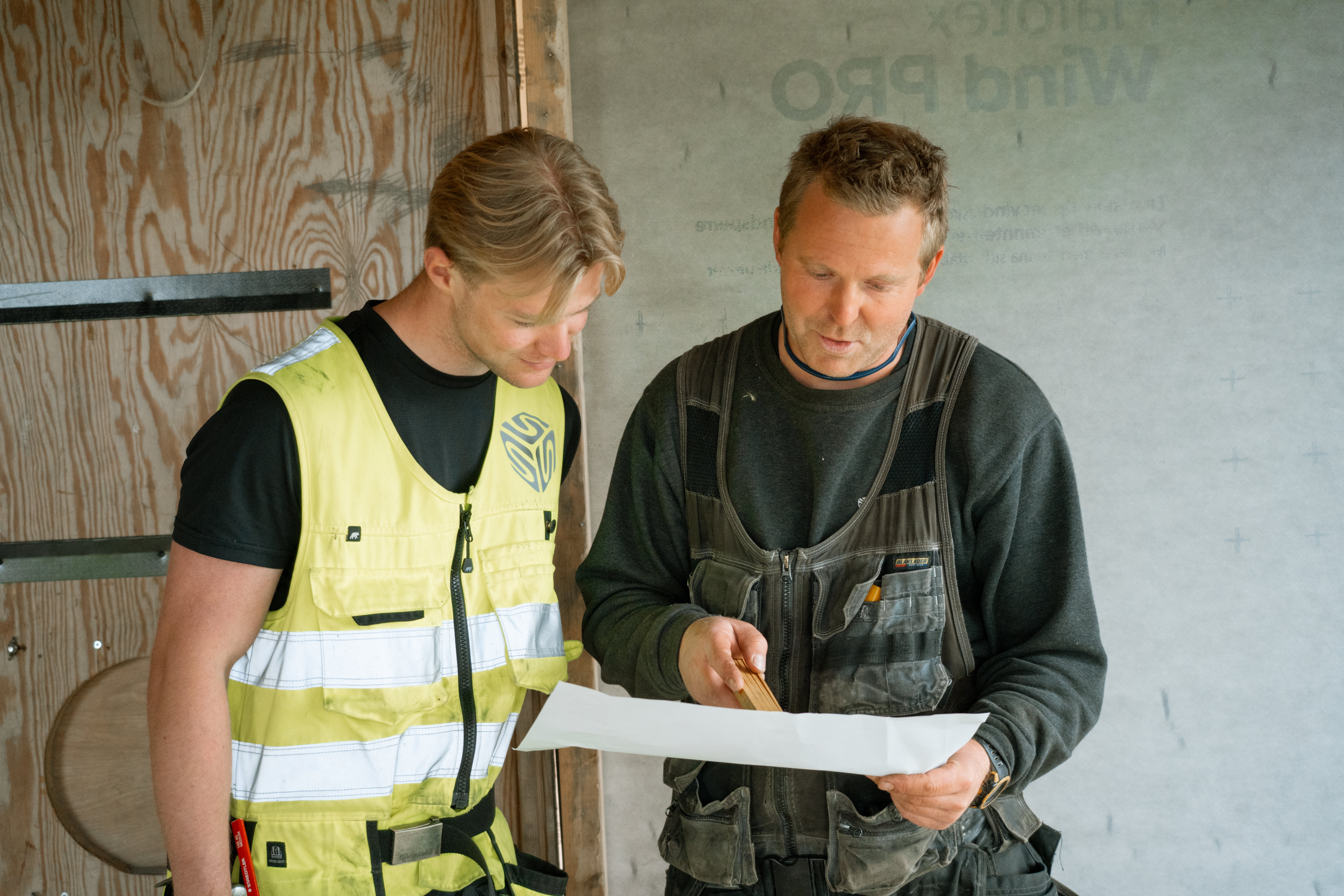To personar i arbeidsklede ser på ei byggeteikning inne på ein byggeplass med treveggar.