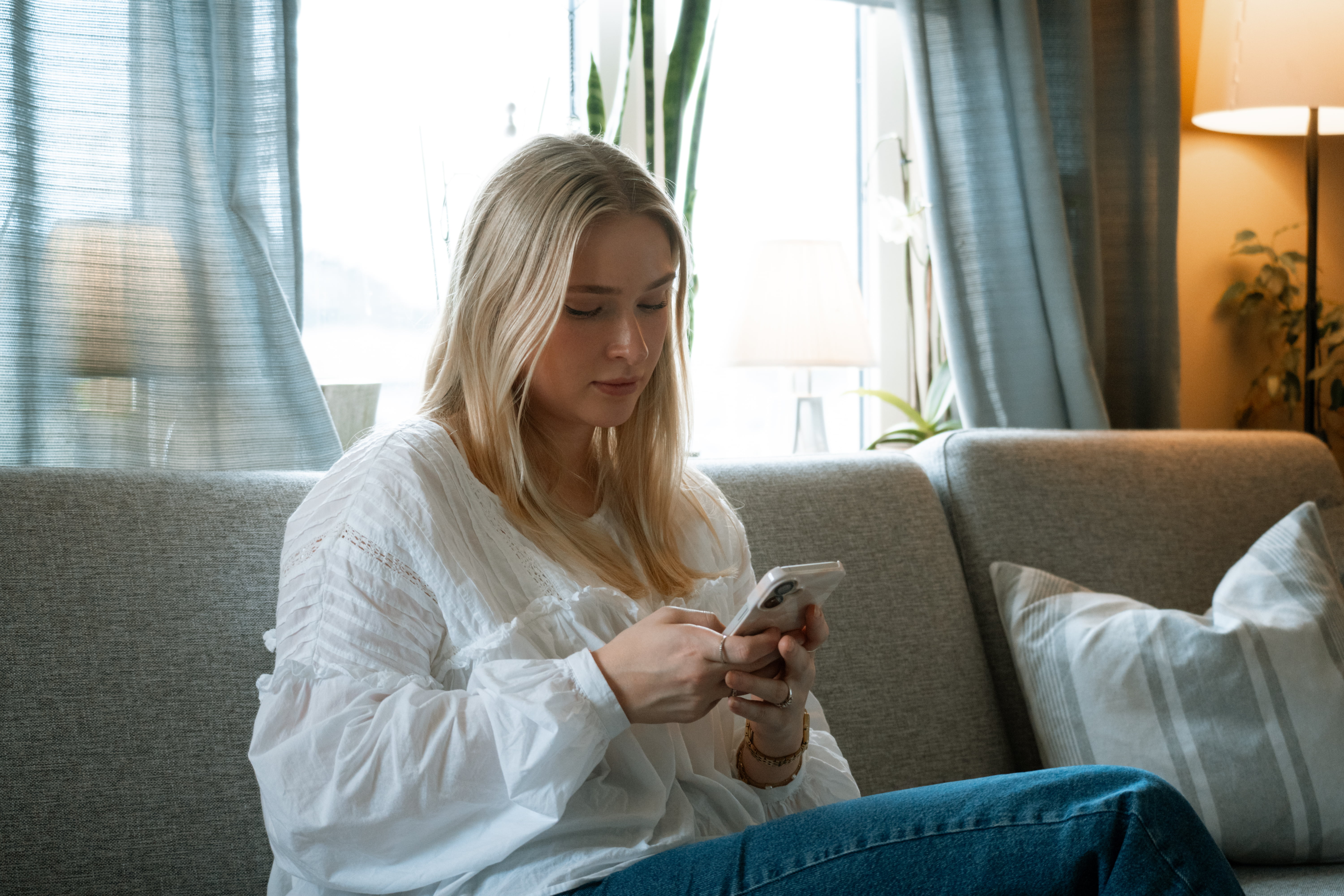 Person med kvit bluse sit i sofaen og held ein mobil, med klokke på handledet.