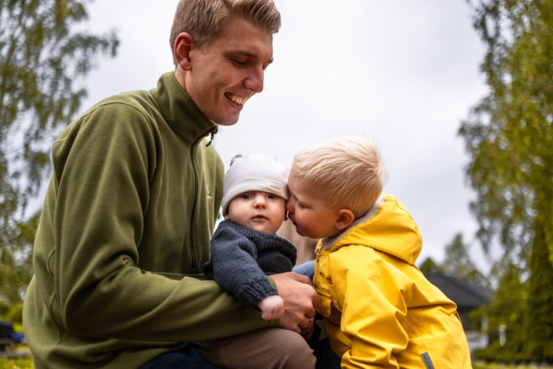 Far med veslebror på fanget sitt og storebror gir han ein klem