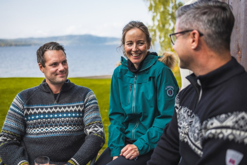 To menn og ei kvinne sitt i hytteveggen framfor eit vatn