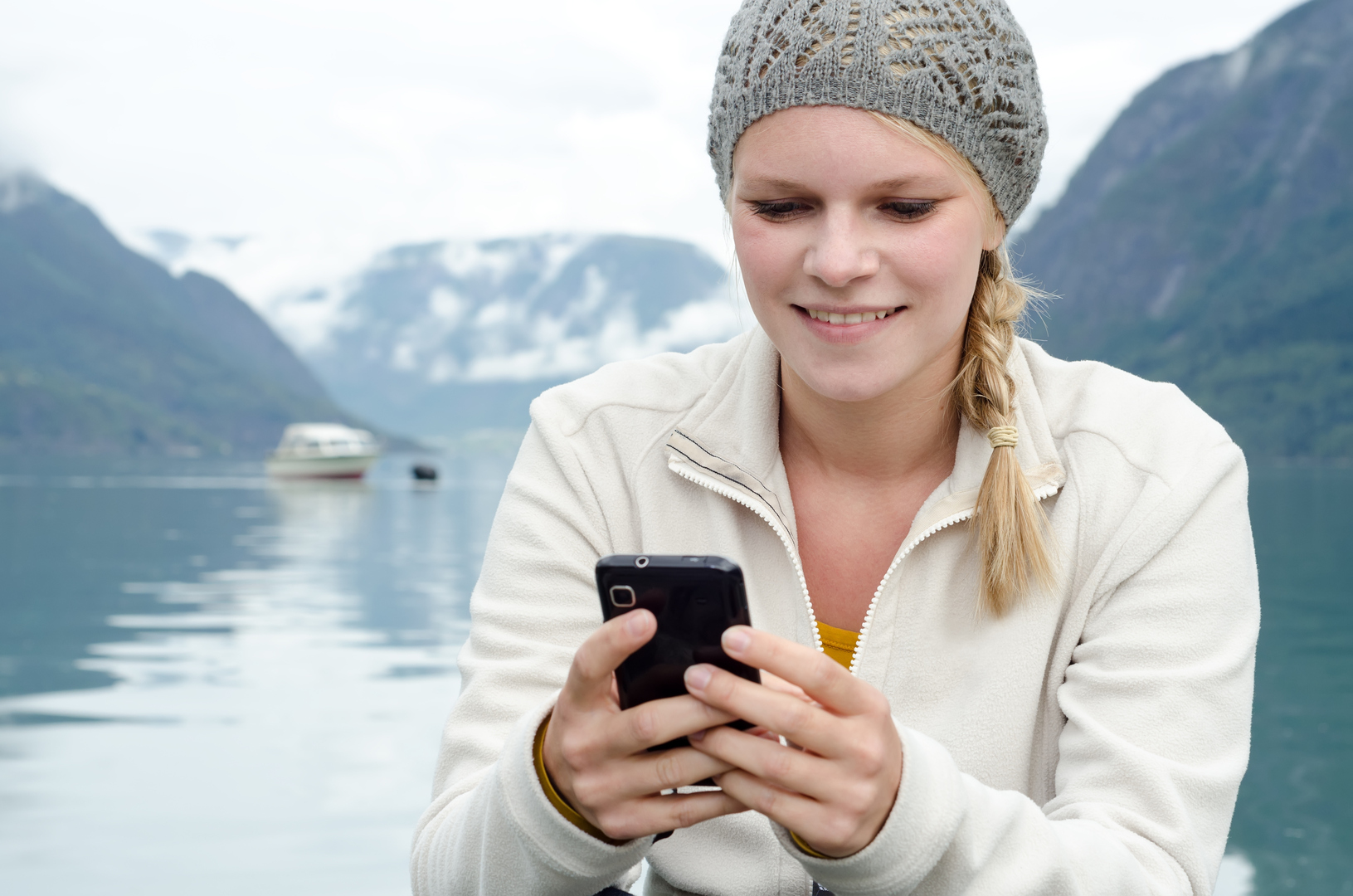 Dame som sitter ved en fjord og ser på mobilen